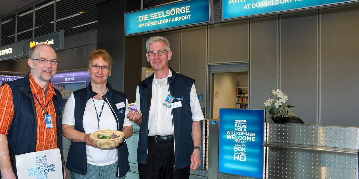 Der Schalter der Flughafenseelsorge in Terminal A, Abflugebene ist gut erkennbar an dem himmelblauen Logo.. Auf dem Bild (v.l.n.r.) sind Pastoralreferent Johannes Westerdick, Diplom-Sozialpädagogin Ute Clevers und der ehrenamtliche Mitarbeiter Jan-Georg Simon.