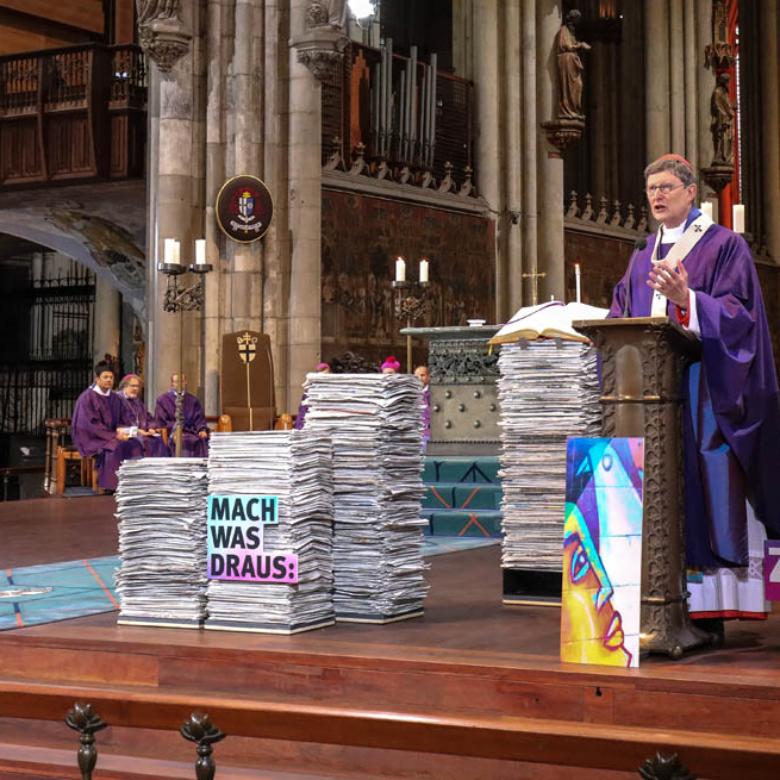 Kardinal Woelki predigt während des Pontifikalamts umgeben von Zeitungsstapeln mit Nachrichten aus aller Welt, die die Frage aufwerfen, wie die Zukunft aussehen soll. Der Erzbischof rief dazu aus aktiv zu werden, um eine gute Zukunft für die Welt zu gestalten.