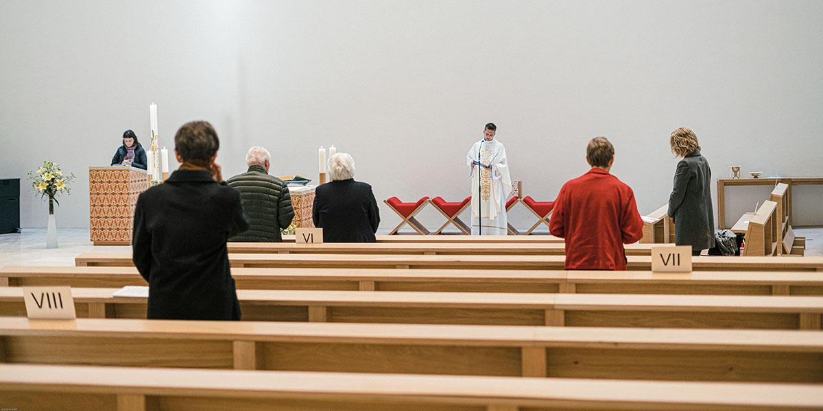 Gottesdienstbesucher stehen nach Anmeldung und mit Abstand zueinander in den Kirchenbänken während einer Messe. Auf den Kirchenbänken platzierte Zettel mit römischen Ziffern markieren mögliche Sitzplätze für Gottesdienstbesucher.