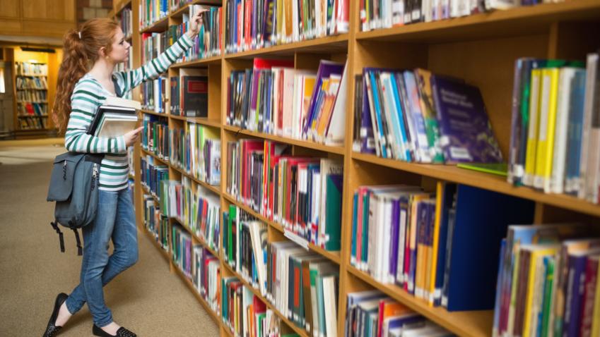 Redhead student taking book from shelf in the library at the university