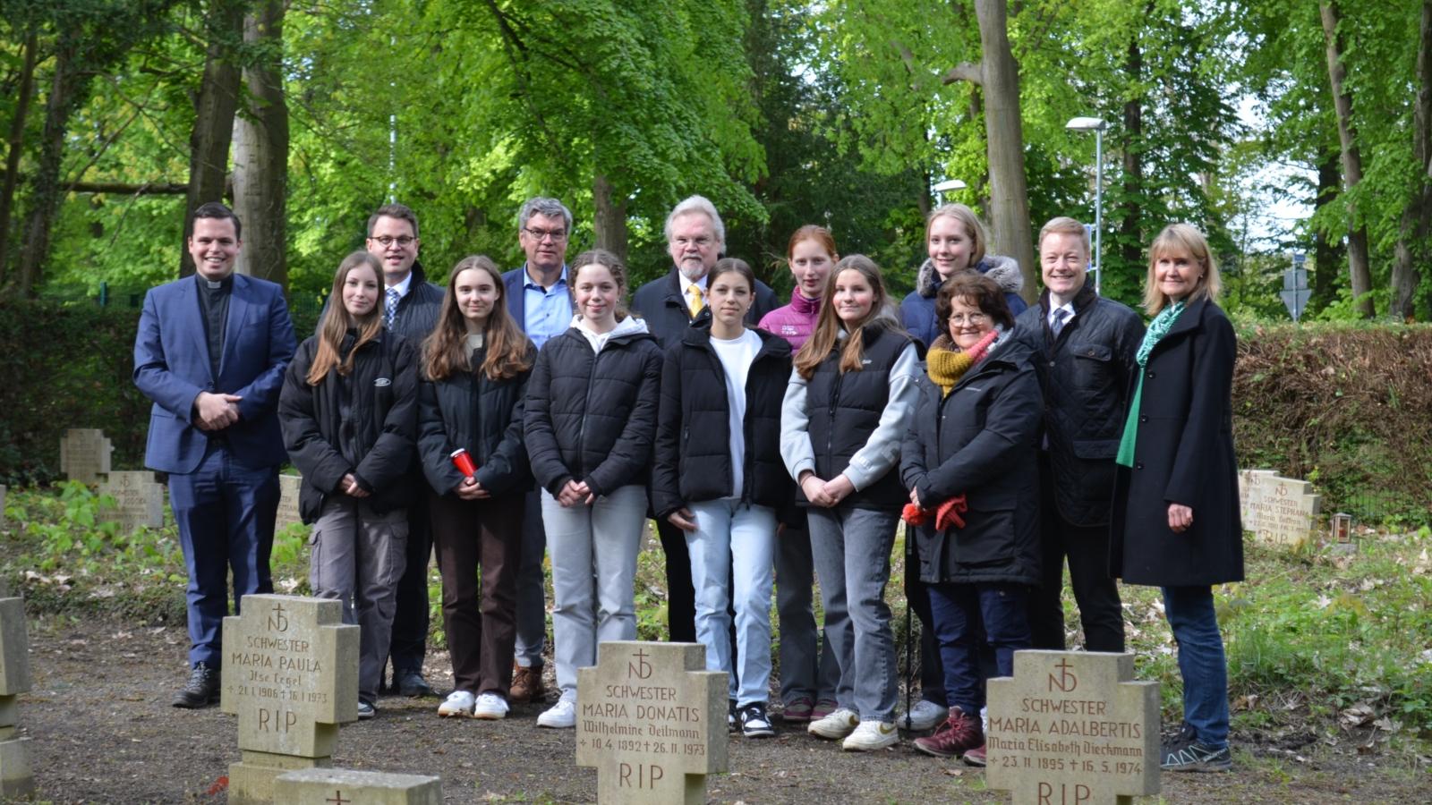 Gruppenbild auf dem Friedhof (v.l.): Daniel Sluminsky (Schulseelsorger), Rafael Bartsch (Erzb. Schulrat), Sophia, Magdalena, Michael Bornemann (Schulleiter), Clara, Norbert Erlinghagen (Vorstand Schulfonds), Olivia, Carlotta, Emma, Josefine, Anna van dem Brink (Religionslehrerin), Thomas Pitsch (Bereichsleiter Schule & Hochschule, Erzbistum Köln), Petra Hennigfeld (stellv. Schulleiterin und Projektbetreuerin).