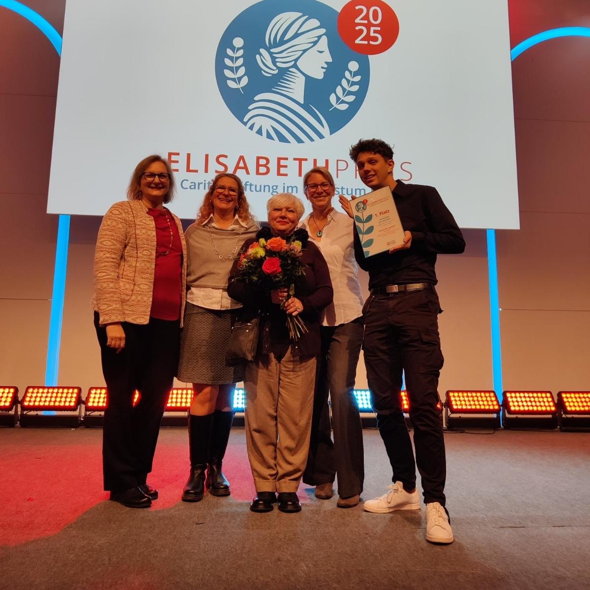 Ein Foto zeigt fünf Personen auf einer Bühne. Im Hintergrund ist das Logo des Elisabeth Preis zu sehen.