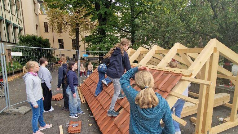 Ein Foto zeigt Schülerinnen die an einem Modelldachstuhl Dachziegeln montieren.