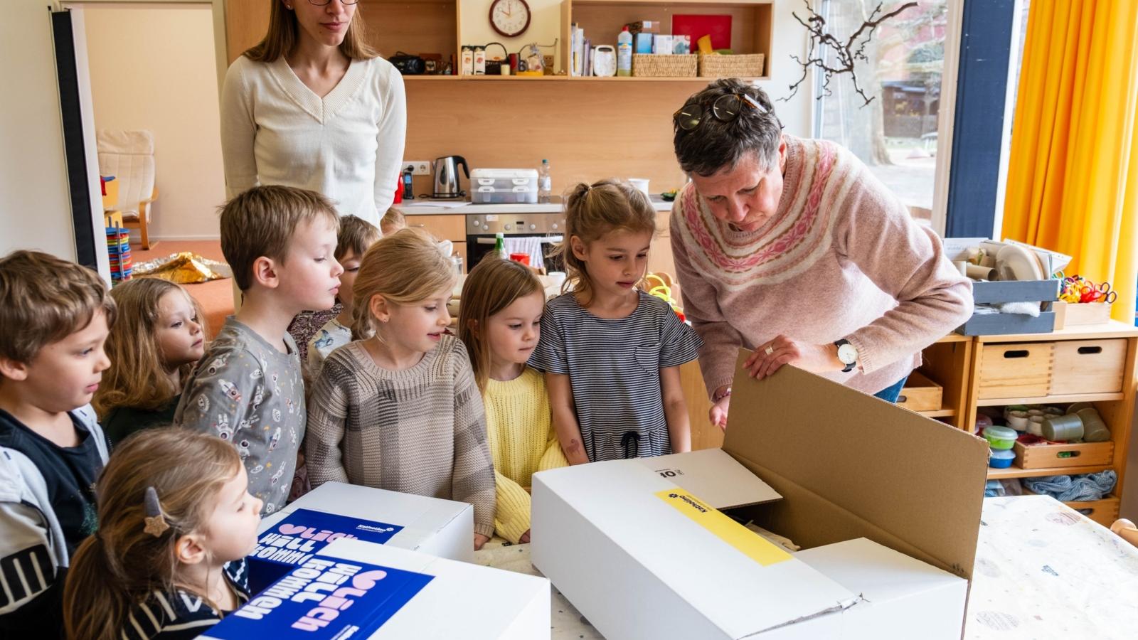 Eine Frau packt ein Paket aus. Um das Paket stehen sieben Kita-Kinder und schauen zu.