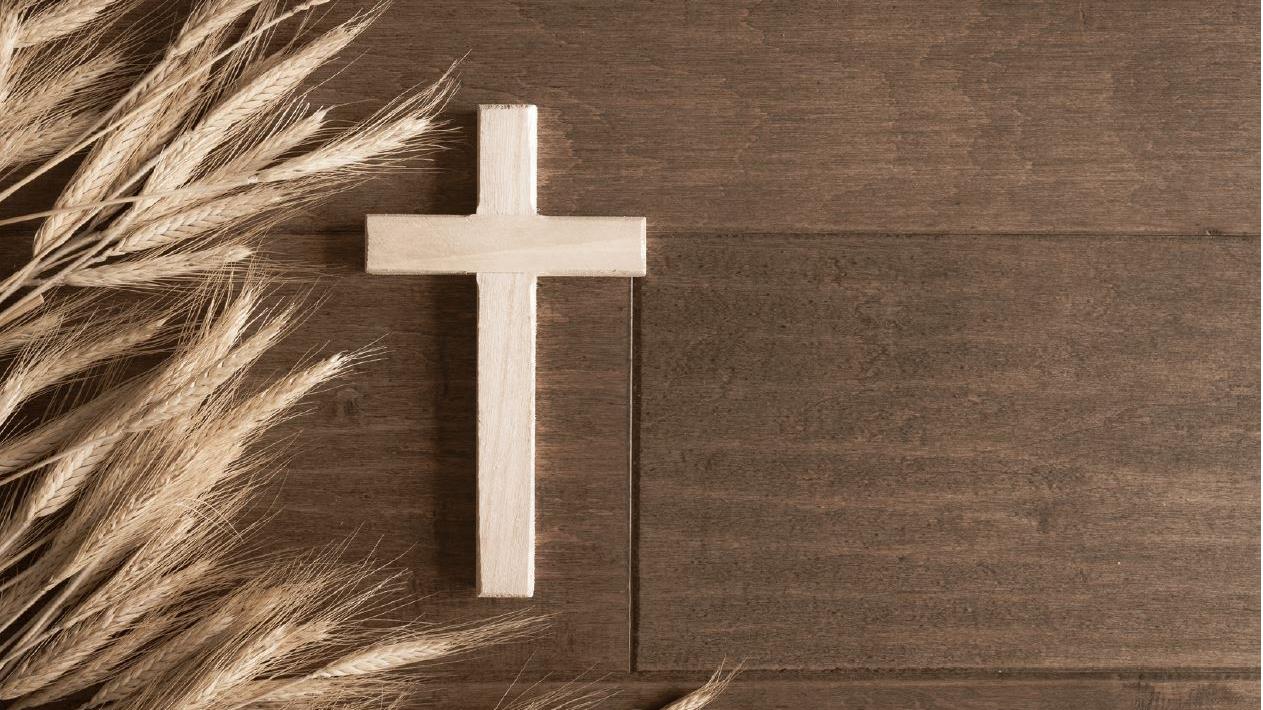 helles Holzkreuz ohne Corpus neben Ähren auf dunklem Holzboden