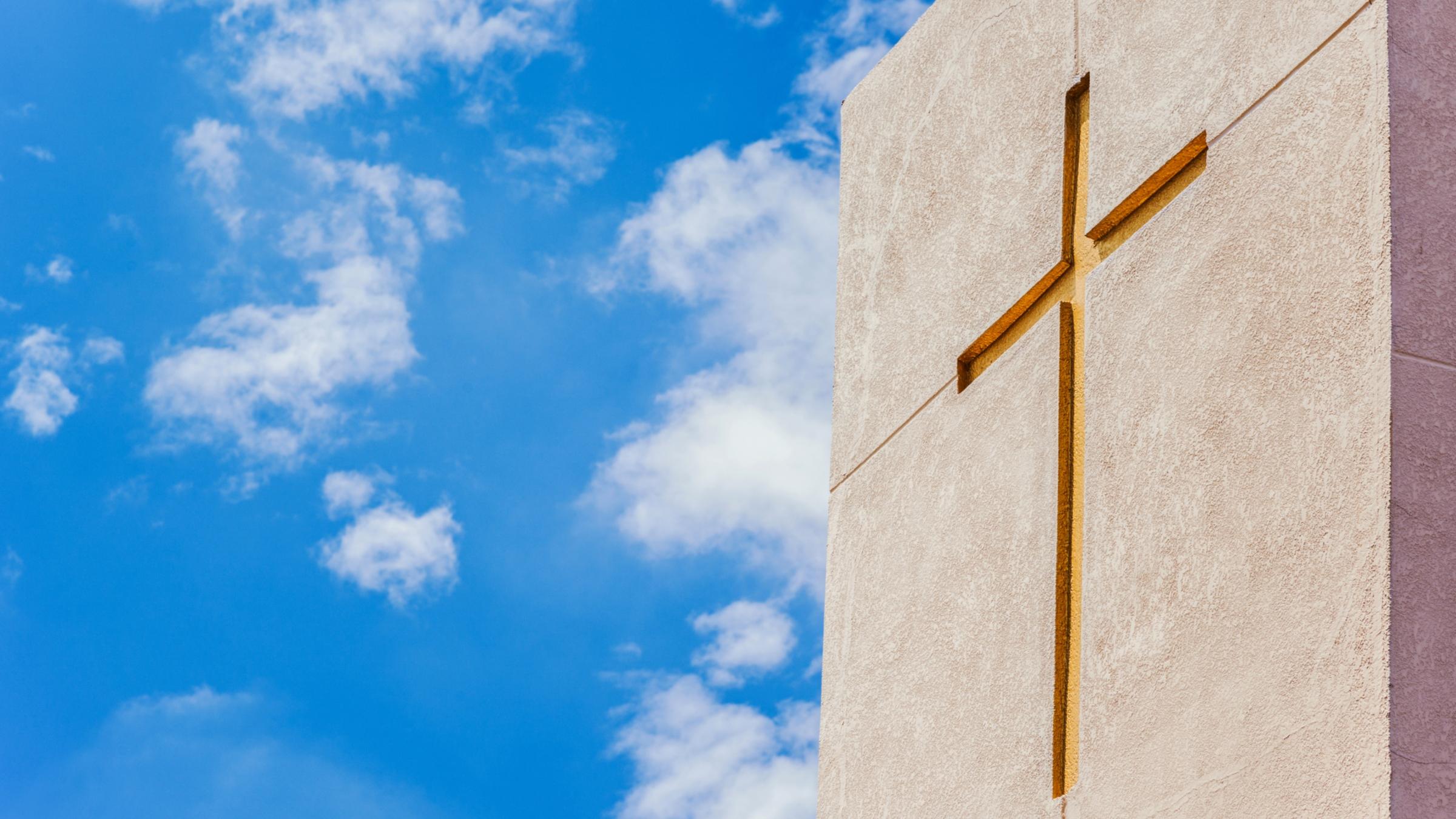 Betonfassade mit eingelassenem goldenen Kreuz, aufgenommen gegen einen leicht bewölkten strahlend blauen Himmel