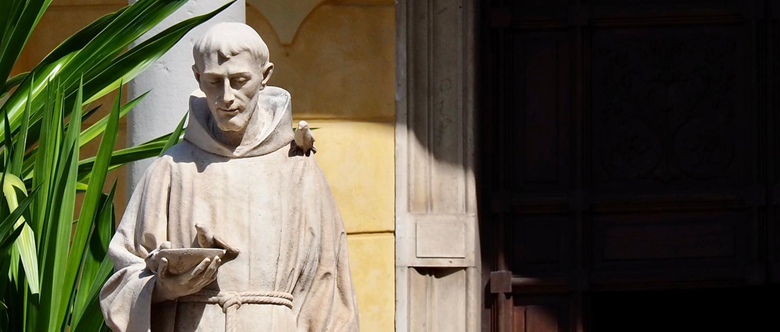 Skulptur des hl. Franziskus auf dem Vorplatz der Chiesa Dei Cappuccini in Sanremo, Italien Steinstatue eines Mönchs mit Buch in sonnigem Hof neben einer Pflanze