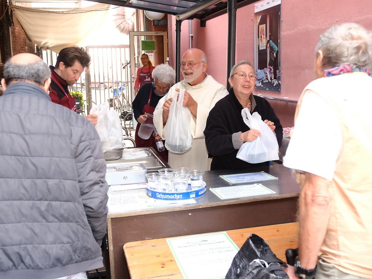 Ein Foto zeigt zwei Personen von hinten vor einem Tresen. Hinter dem Tresen stehen ein Mann und eine Frau die Tüten austeilen.