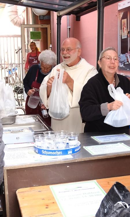 Ein Foto zeigt zwei Personen von hinten vor einem Tresen. Hinter dem Tresen stehen ein Mann und eine Frau die Tüten austeilen.