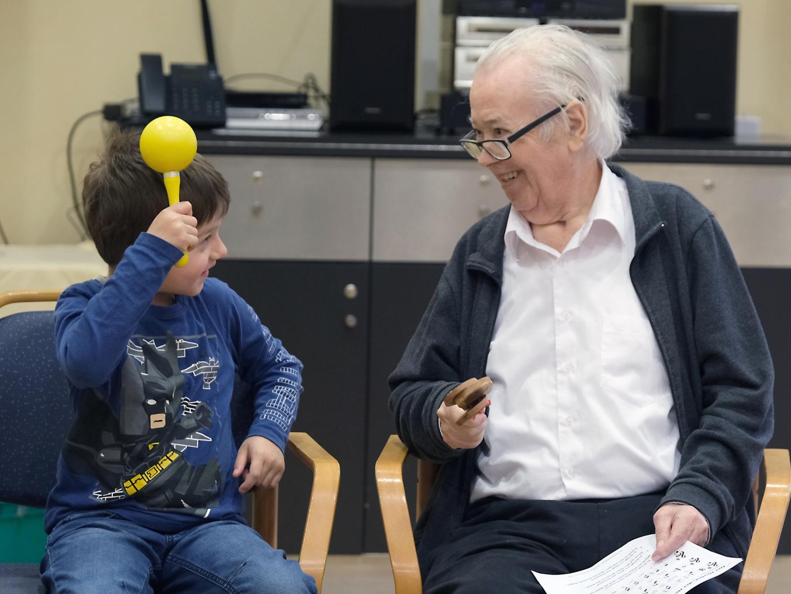 Ein Foto zeigt ein Kind und einen alten Mann mit einer Rassel auf Stühlen sitzend.