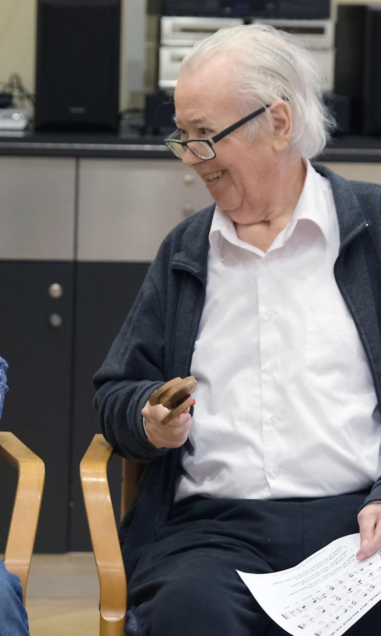 Ein Foto zeigt ein Kind und einen alten Mann mit einer Rassel auf Stühlen sitzend.