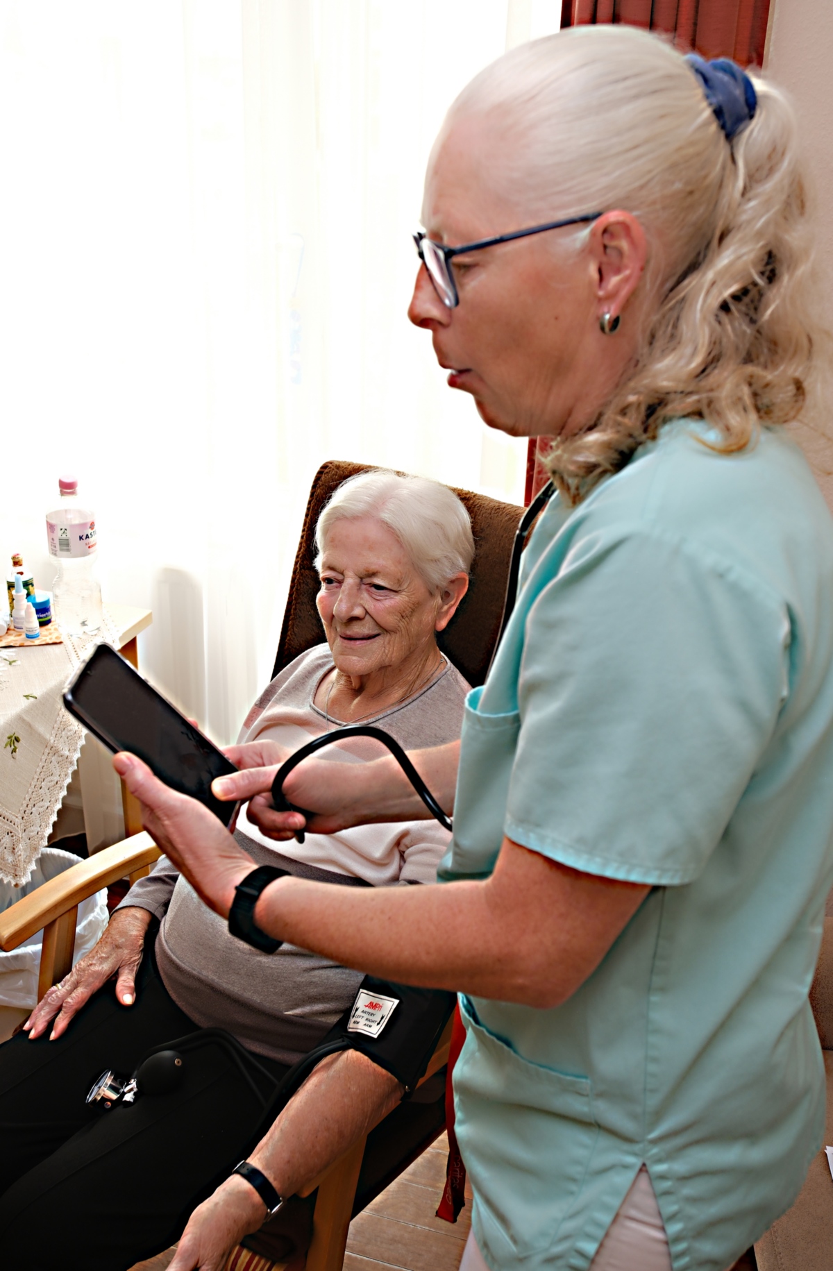 Ein Foto zeigt eine Frau mit Handy und  in der Hand. Im Hintergrund sitzt eine ältere Frau in einem Sessel.