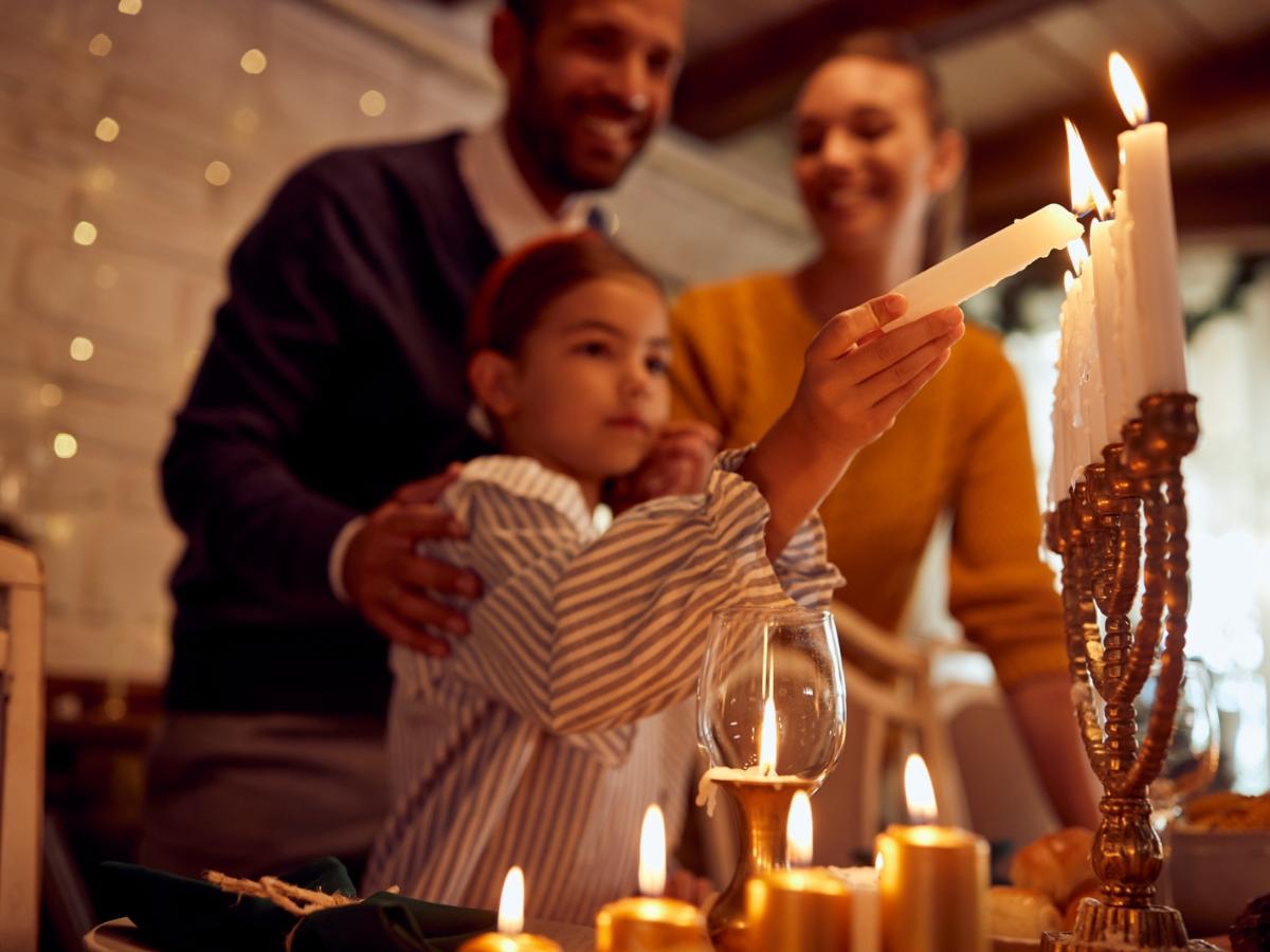 Ein Foto zeigt ein Mädchen und hinter ihr zwei Erwachsene. Das Mädchen zündet mit einer Kerze weitere Kerzen auf einem Leuchter an.