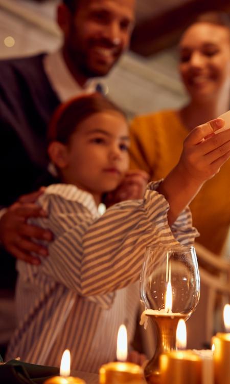 Ein Foto zeigt ein Mädchen und hinter ihr zwei Erwachsene. Das Mädchen zündet mit einer Kerze weitere Kerzen auf einem Leuchter an.