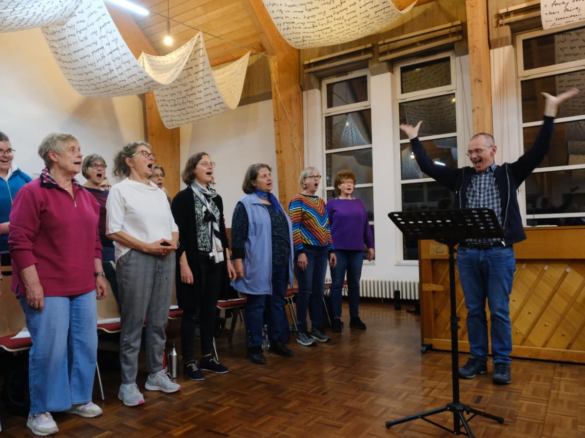 Ein Foto zeigt einen Mann mit erhobenen Armen auf der rechten Seite und mehrere singenden Personen auf der linken Seite.