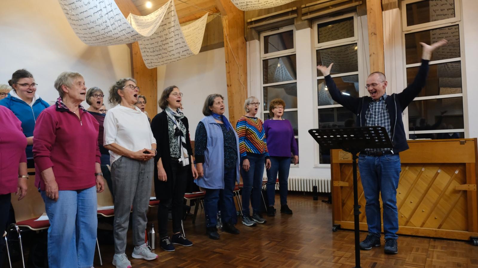 Ein Foto zeigt einen Mann mit erhobenen Armen auf der rechten Seite und mehrere singenden Personen auf der linken Seite.