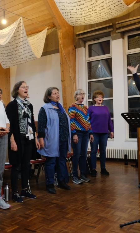 Ein Foto zeigt einen Mann mit erhobenen Armen auf der rechten Seite und mehrere singenden Personen auf der linken Seite.